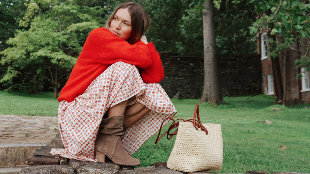 A woman sits on a stone bench in a park-like setting, wrapped in a bright red knit sweater and wearing a white dress with a red geometric pattern. A cream woven tote bag with brown handles rests beside her on the bench.